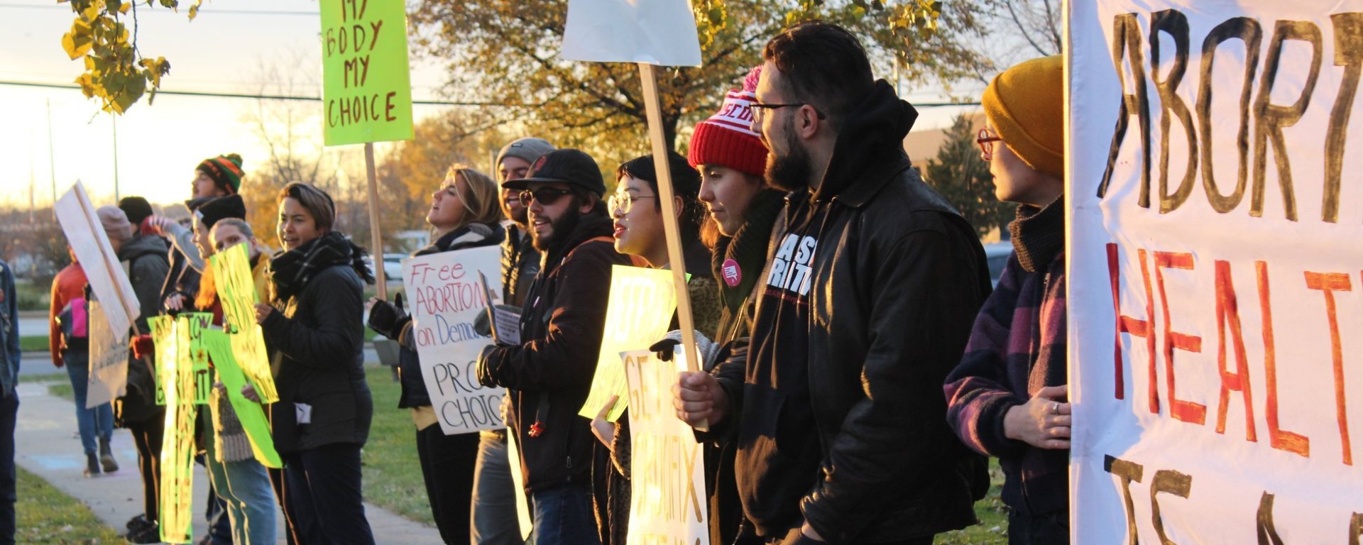 A picture of people standing in a line, holding signs in support of abortion rights, while the sun sets.