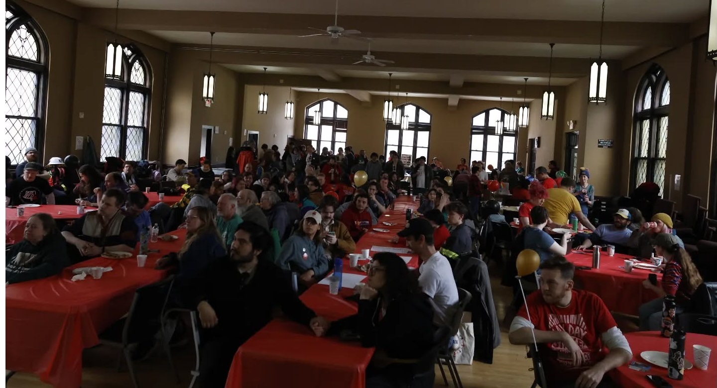 Image shows several rows of tables in a dimly lit non-denominational church space, filled with people listening to a speaker who is outside of the shot.