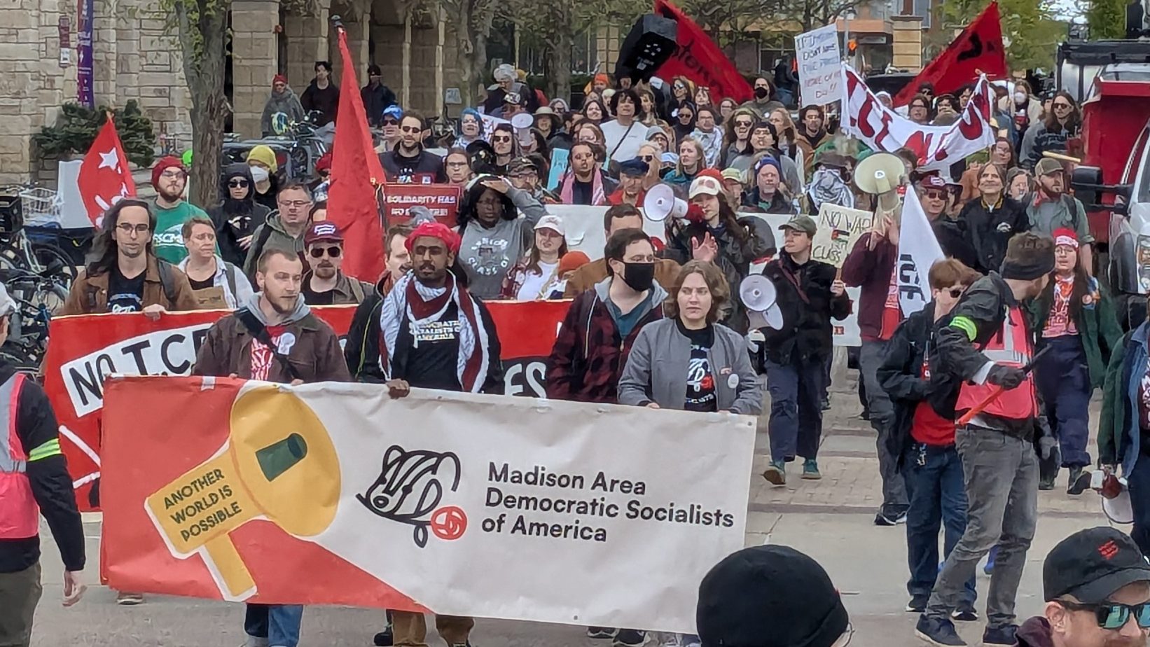 The image shows protesters marching in State Street. Three people at the front of the group hold a wide banner that says Madison Area Democratic Socialists of America.