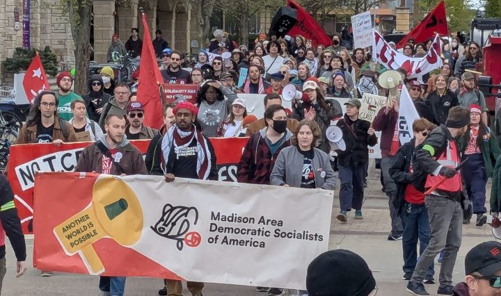 The image shows protesters marching in State Street. Three people at the front of the group hold a wide banner that says Madison Area Democratic Socialists of America.