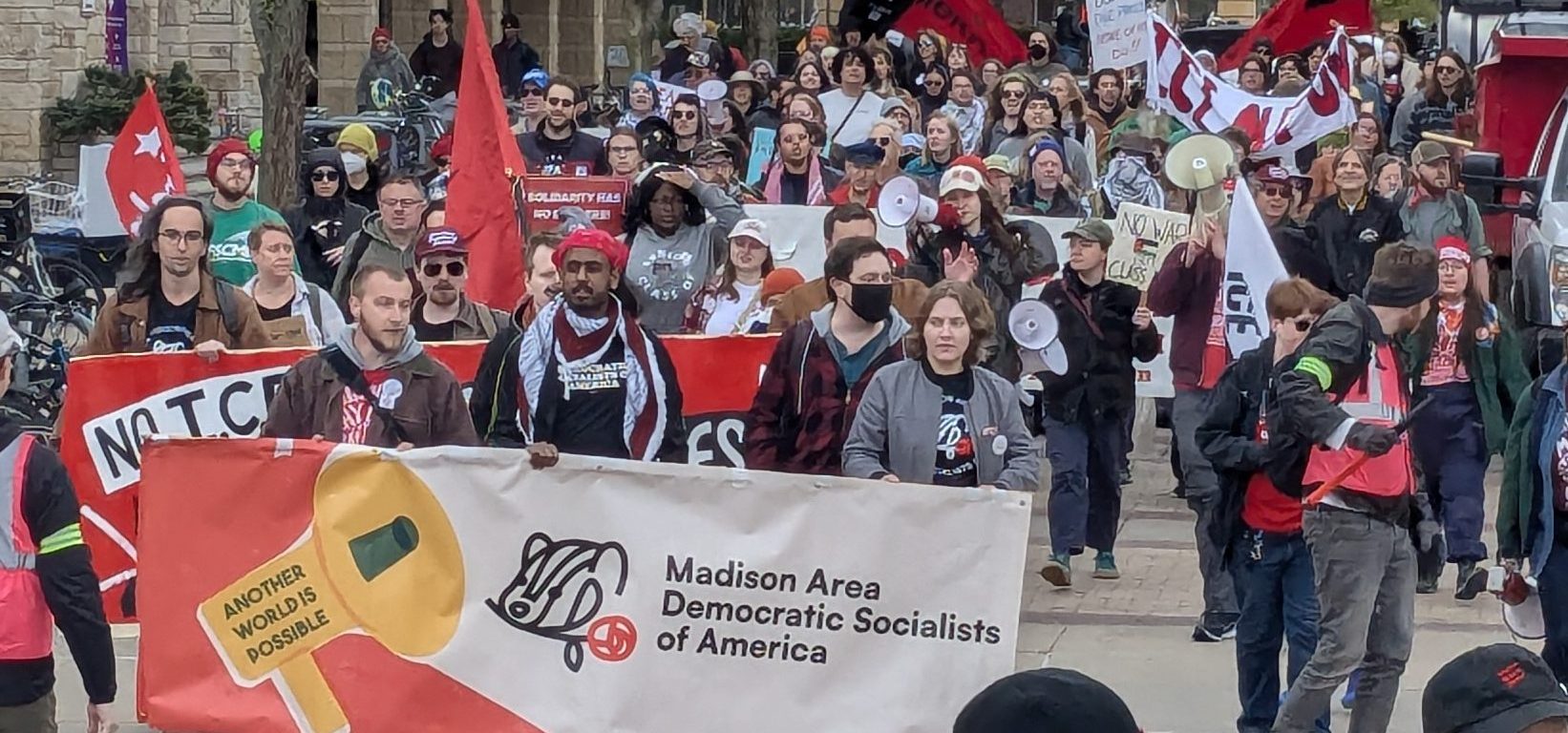 The image shows protesters marching in State Street. Three people at the front of the group hold a wide banner that says Madison Area Democratic Socialists of America.