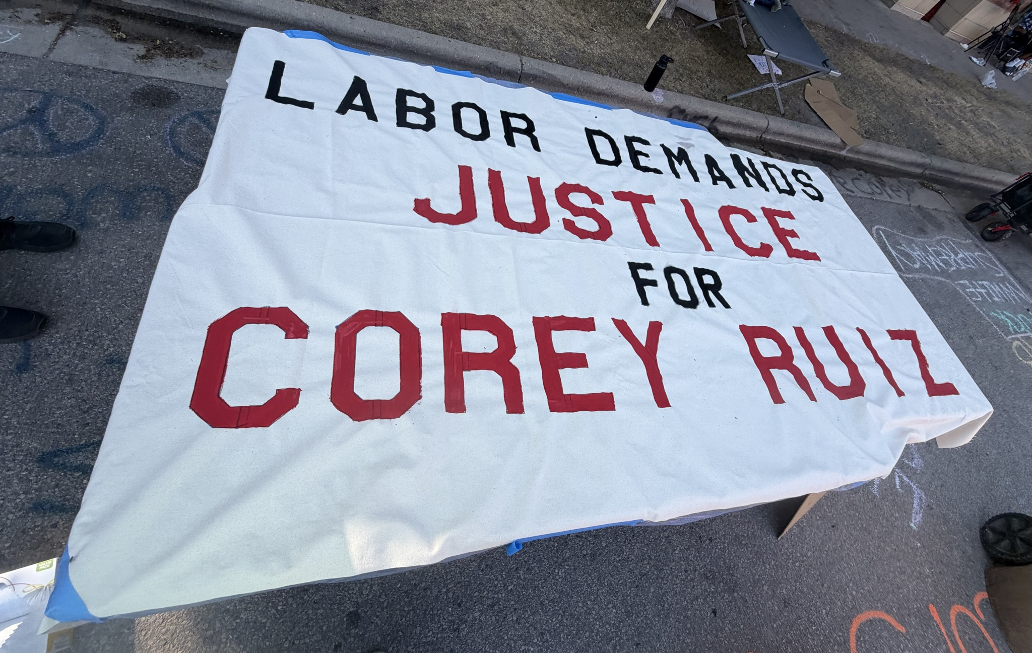 A white poster spread out on the street, with plain text "LABOR DEMANDS JUSTICE FOR COREY RUIZ"